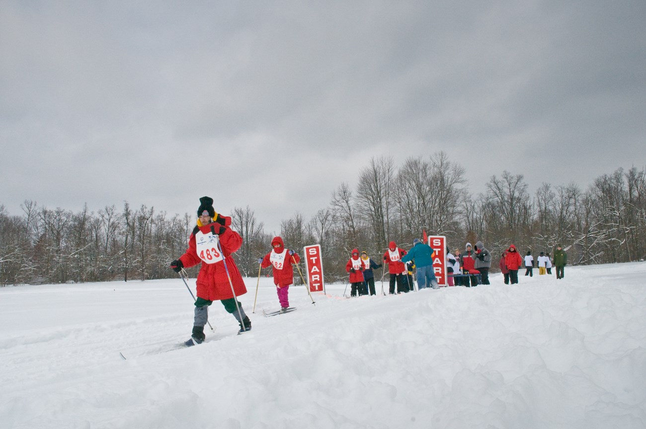 Skiers in red coats and white bibs race from the “Start” line across a flat, snowy field with bare trees beyond.