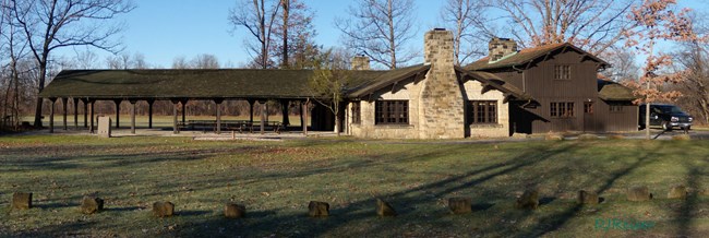 Side view of a gray stone and brown wood picnic shelter, with long, covered porch and stone chimney.