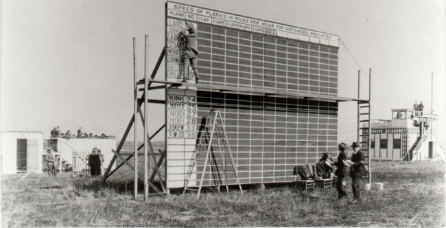 Black and white image of The official scoreboard with scorer updating with racers for  Event No. 2, the National Cash Register Trophy race.