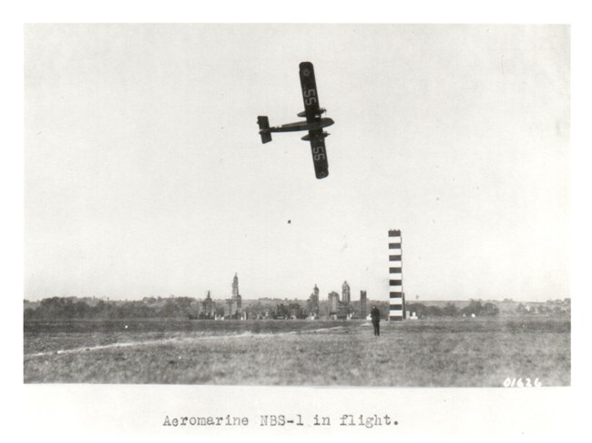 Black and white image of a Aeromarine NBS-1 flying past a pylon on the race course . In the  background can be seen a mock-up of New York City, used as a bombing demonstration on the third day.