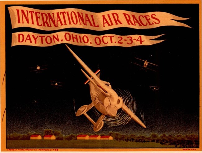 Colored advertisement with the words "International Air Races Dayton, Ohio. October 2-3-4". Cartoonized airplanes appear in the center of the ad. Small houses with red roofs appear along the bottom of the image.