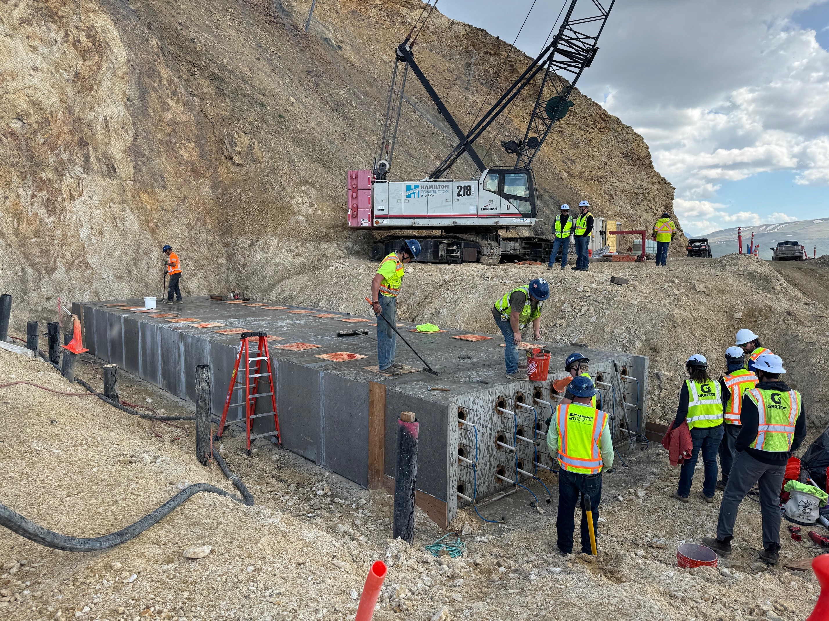 Several construction workers stand on and around and large rectangular abutment