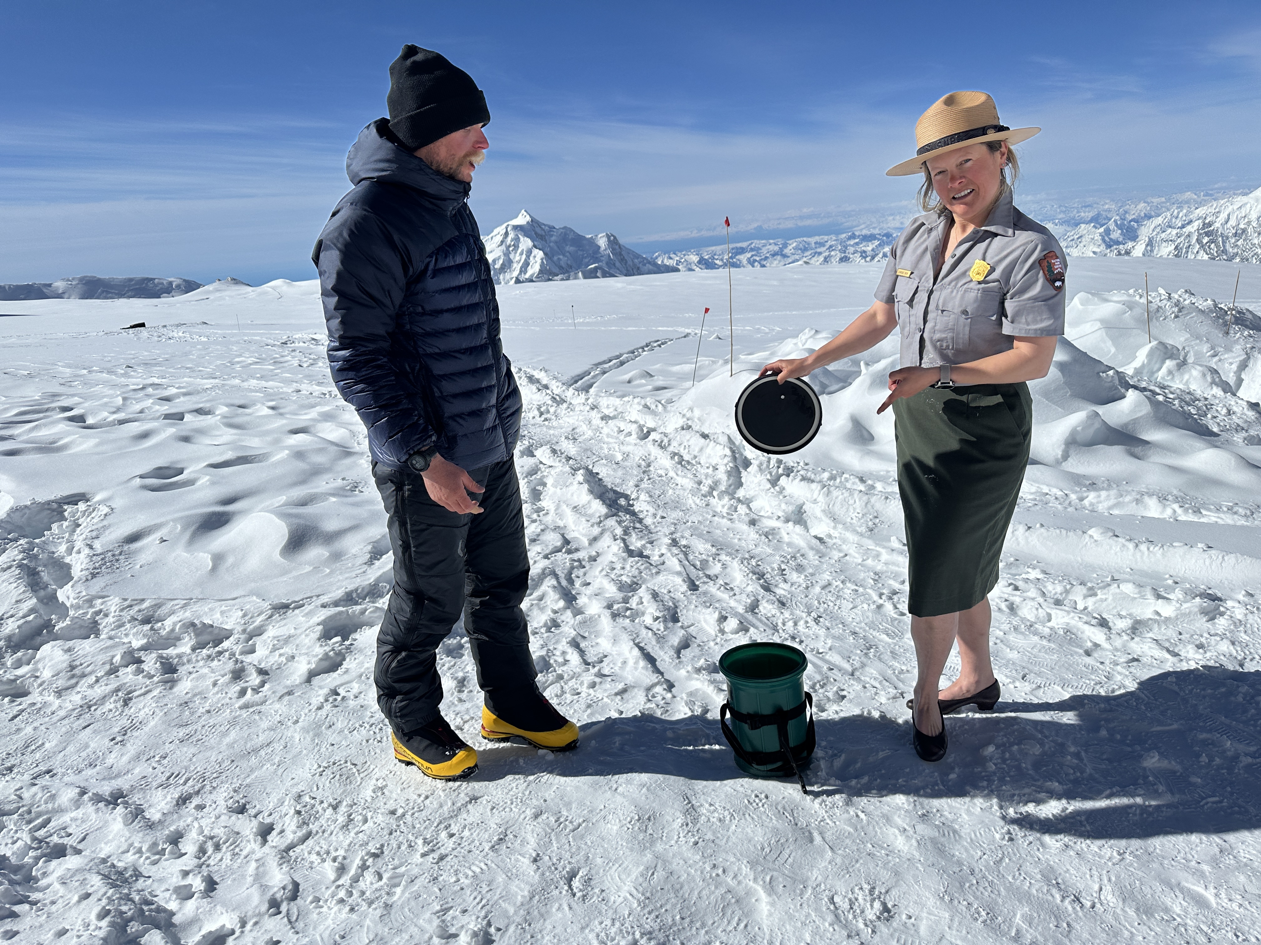 A woman stands on a glacier wearing a NPS park ranger uniform, flat hat, and pumps explains how to use a CMC