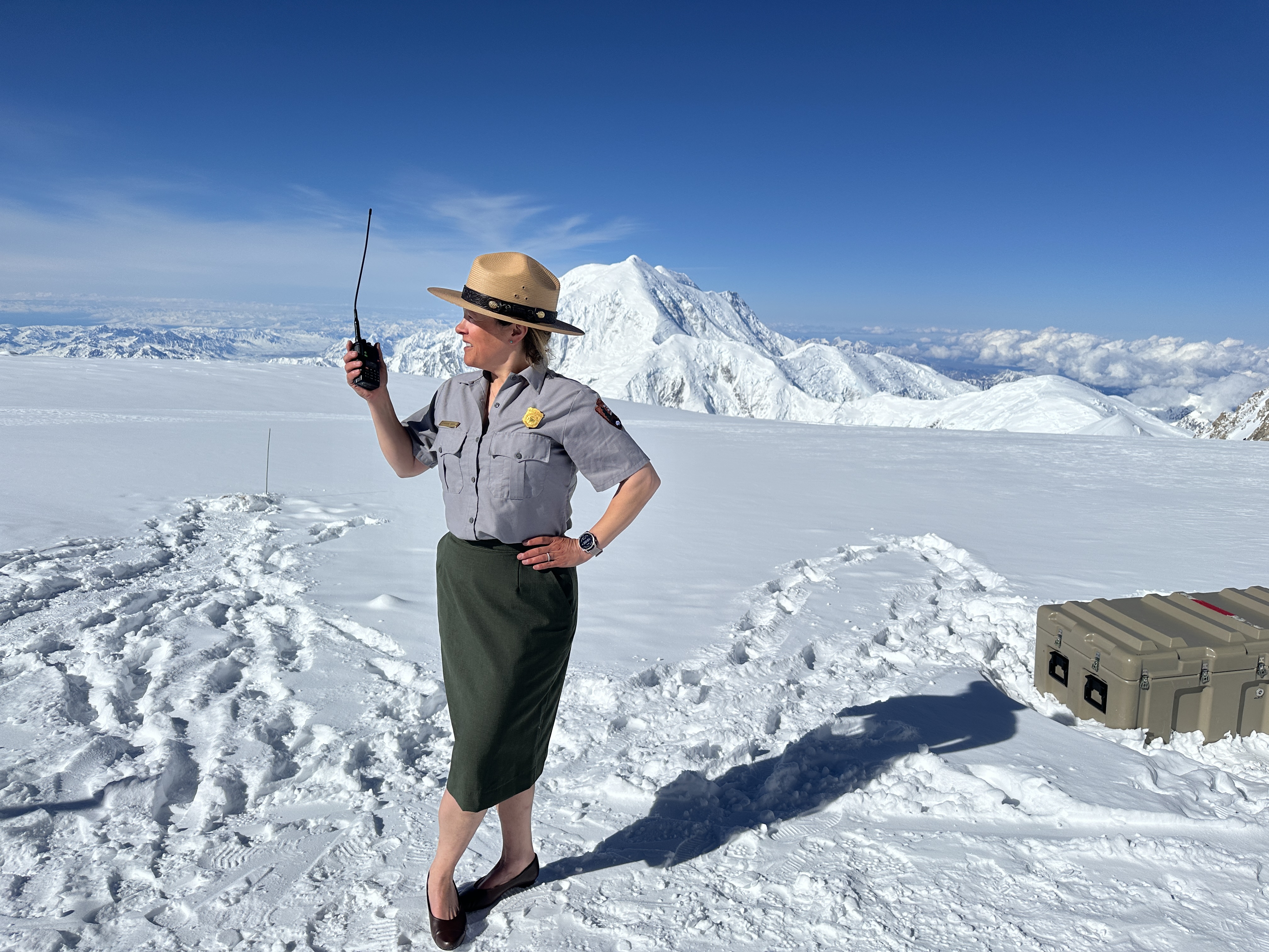 A park ranger in the traditional NPS uniform including pumps, a skirt, and a flat hat, makes a radio call from a glacier