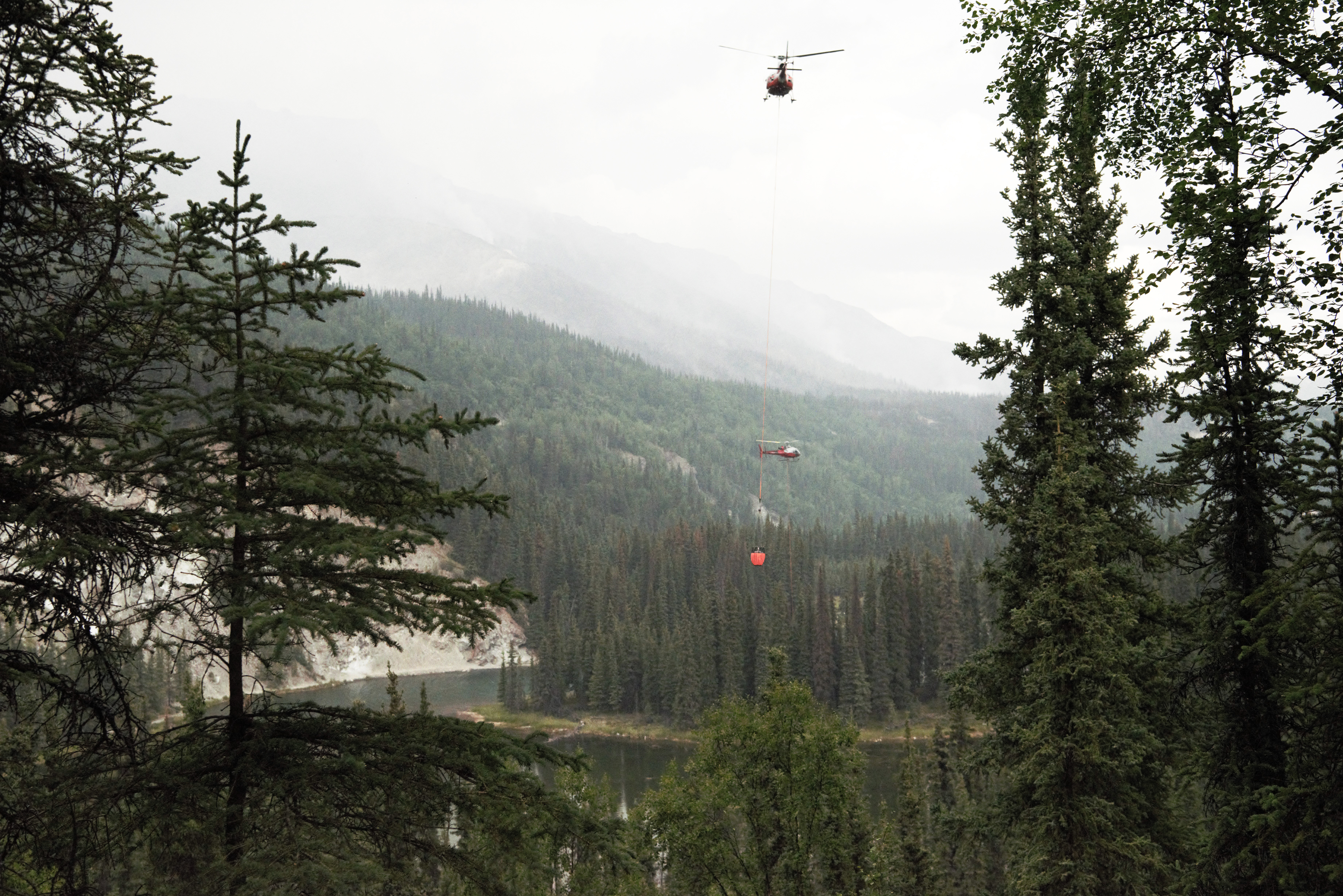 Two red helicopters drawing water from a lake below using orange buckets.  Smoke is rising in the mountainous background