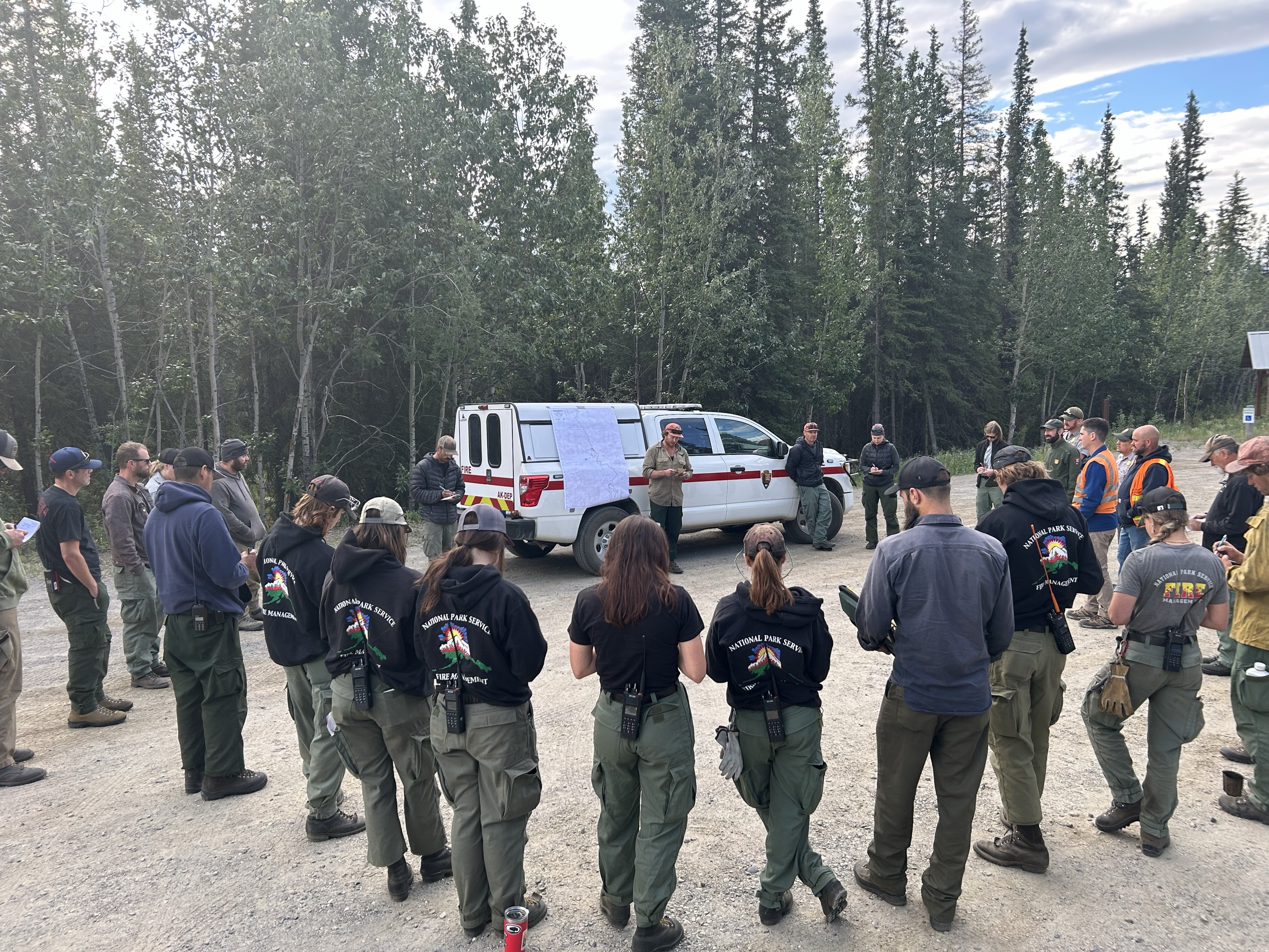 A group of people dressed in fire response clothing, standing in a circle around a vehicle with a map hung on it. The people are taking notes and listening to one presenter.