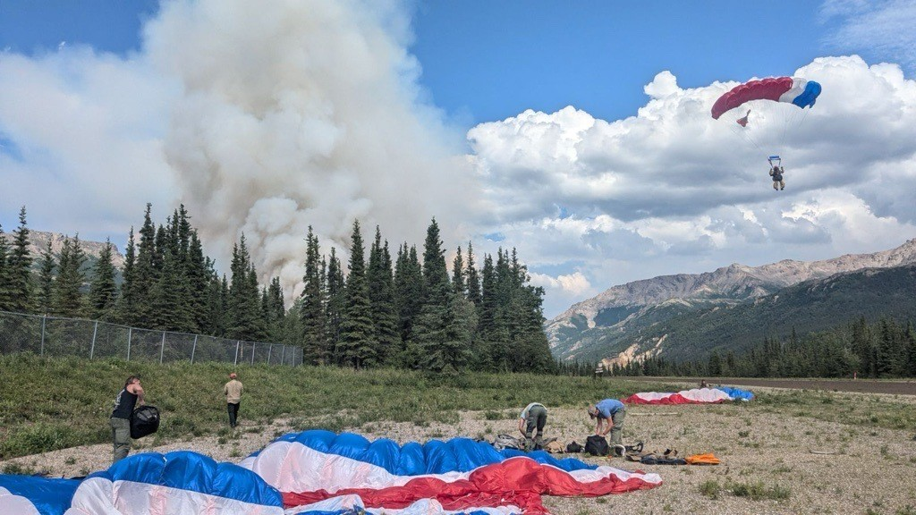 One person descends to the ground via parachute, while four others are surrounded by gear on the ground.