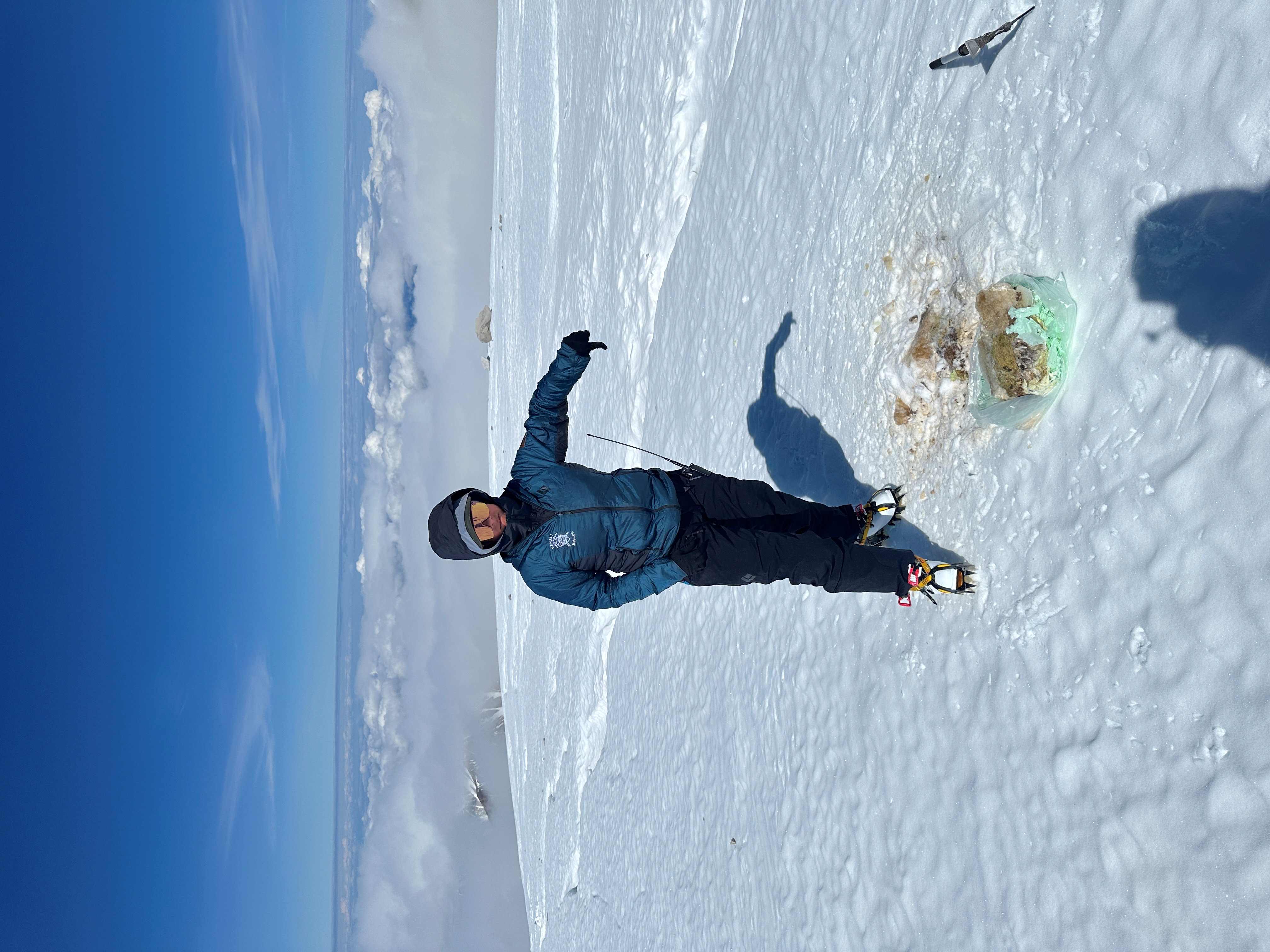 A ranger in a puffy jacket stands on a glacier displaying a big 'thumbs down' over a dirty pile on the glacier