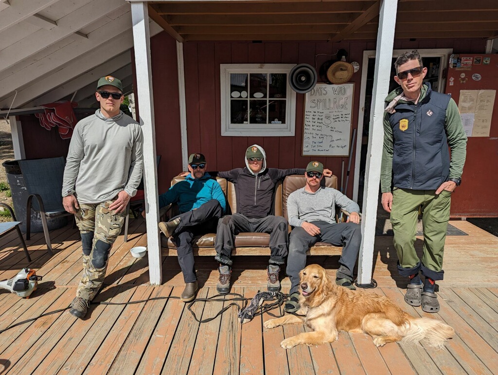 Five men in sunglasses stand on a cabin porch wth a golden retriever