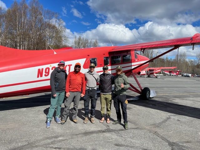 Five climbers stand outside a red and white airplane in street clothes