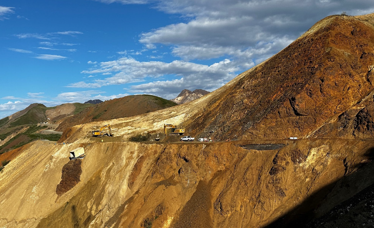 Construction equipment digs along a steep cliff of colorful rocks with a road cut through it.