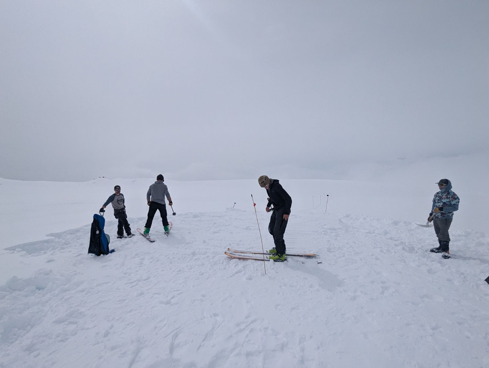 Four patrol members, two on wide skis and two with shovels, prepare a helicopter landing pad on a glacier