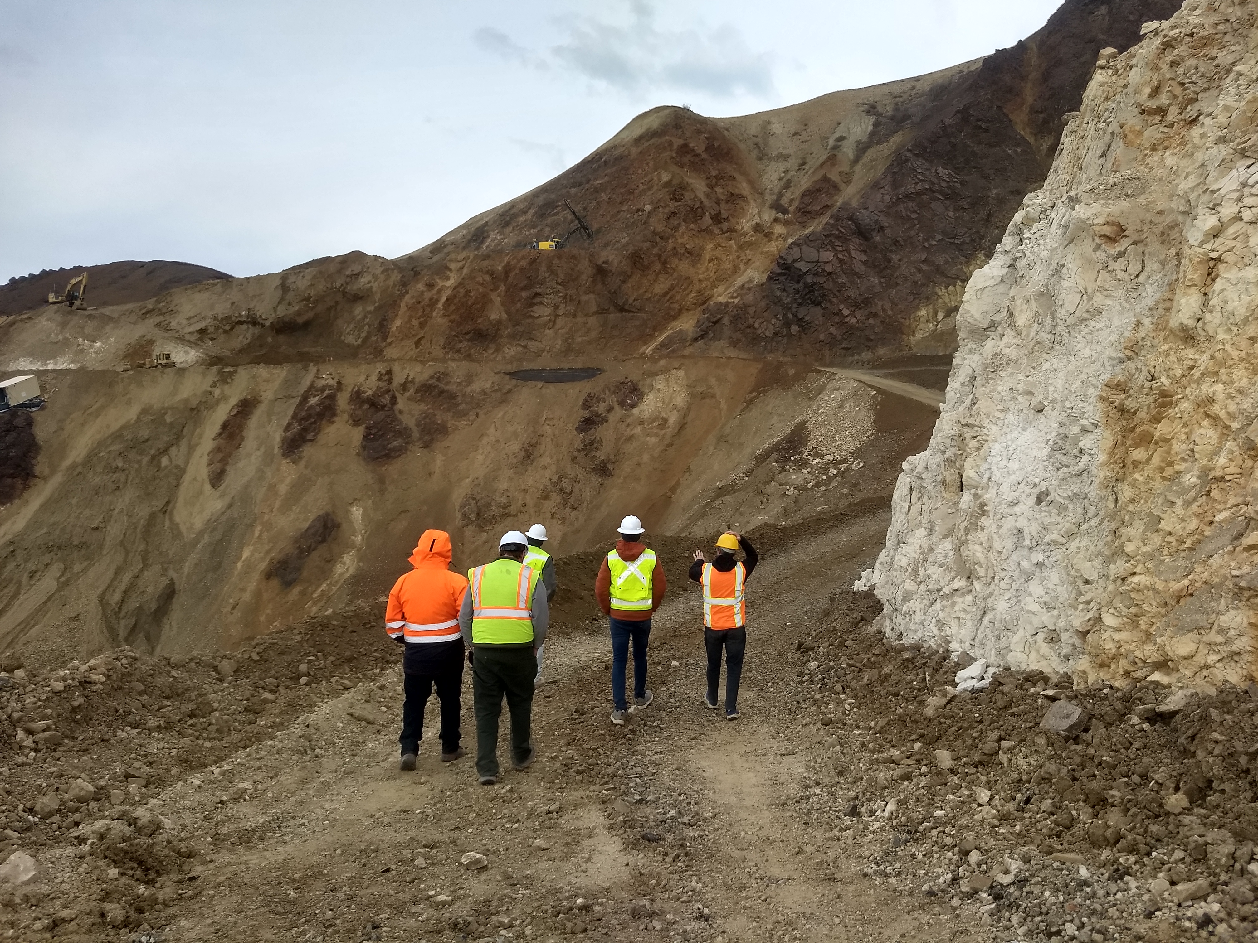 Five people in safety vests and hard hats walk toward the west side of the landslide where several pieces of construction equipment can be seen working.