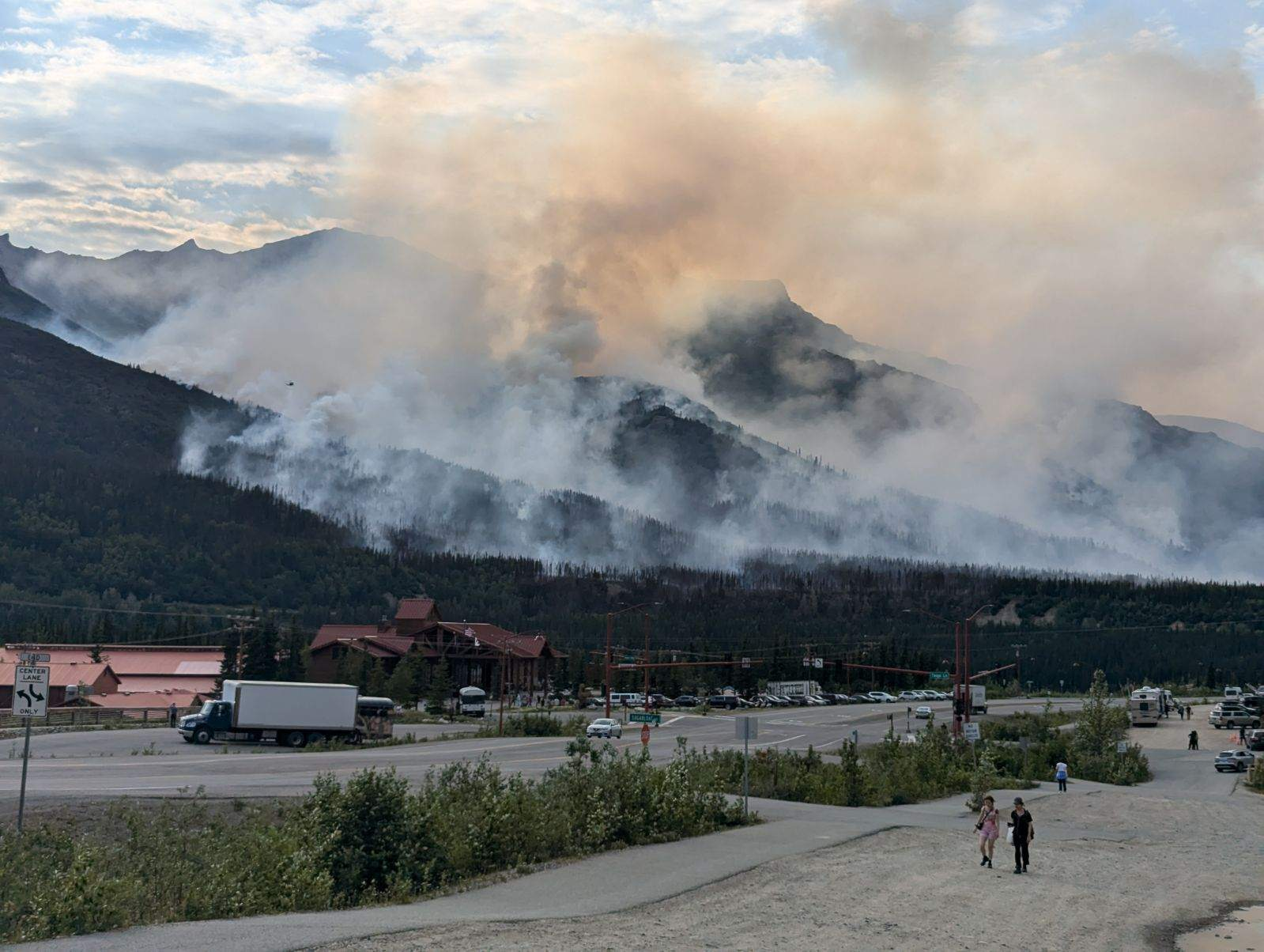 Wildfire smoke rises from forested mountains near a commercial area with buildings, roads, and pedestrians.