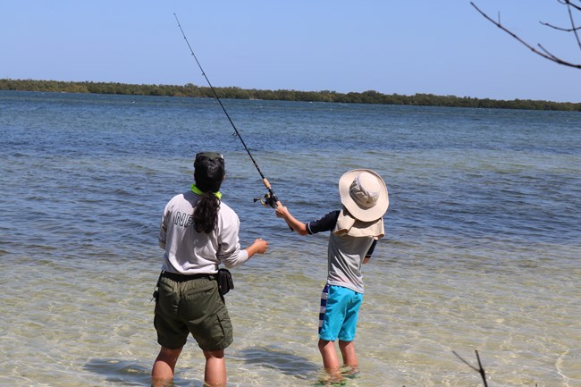 Ranger in green and gray helping a child cast a fishing pole. They are standing in clear water with a clear blue sky in the background.