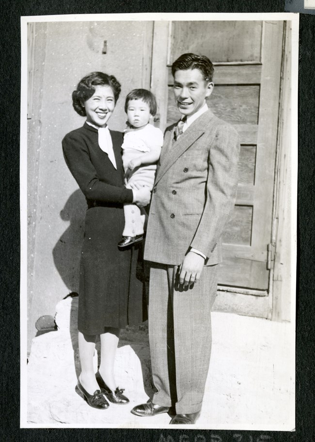 A smiling couple dressed in suits hold their child close, standing in front of an exterior door.