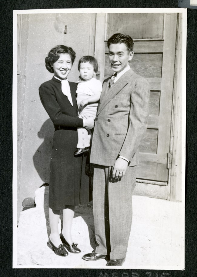 A smiling couple dressed in suits hold their child close, standing in front of an exterior door. A smiling couple dressed in suits hold their child close, standing in front of an exterior door.