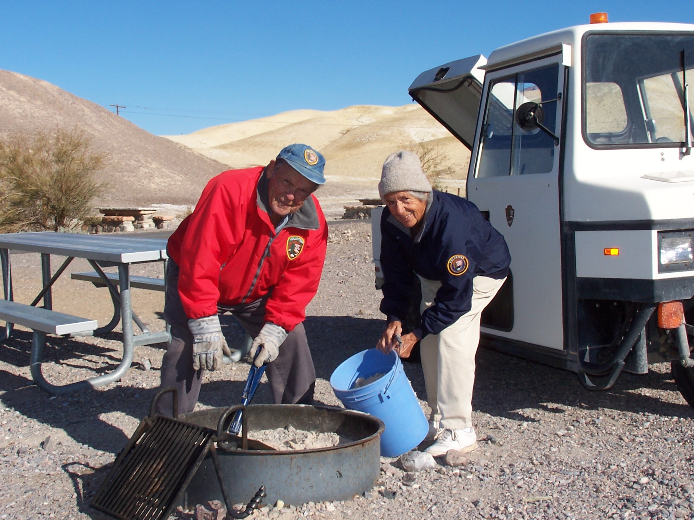 Two volunteers clean ashes out of a fire pit in Texas Springs Campground.