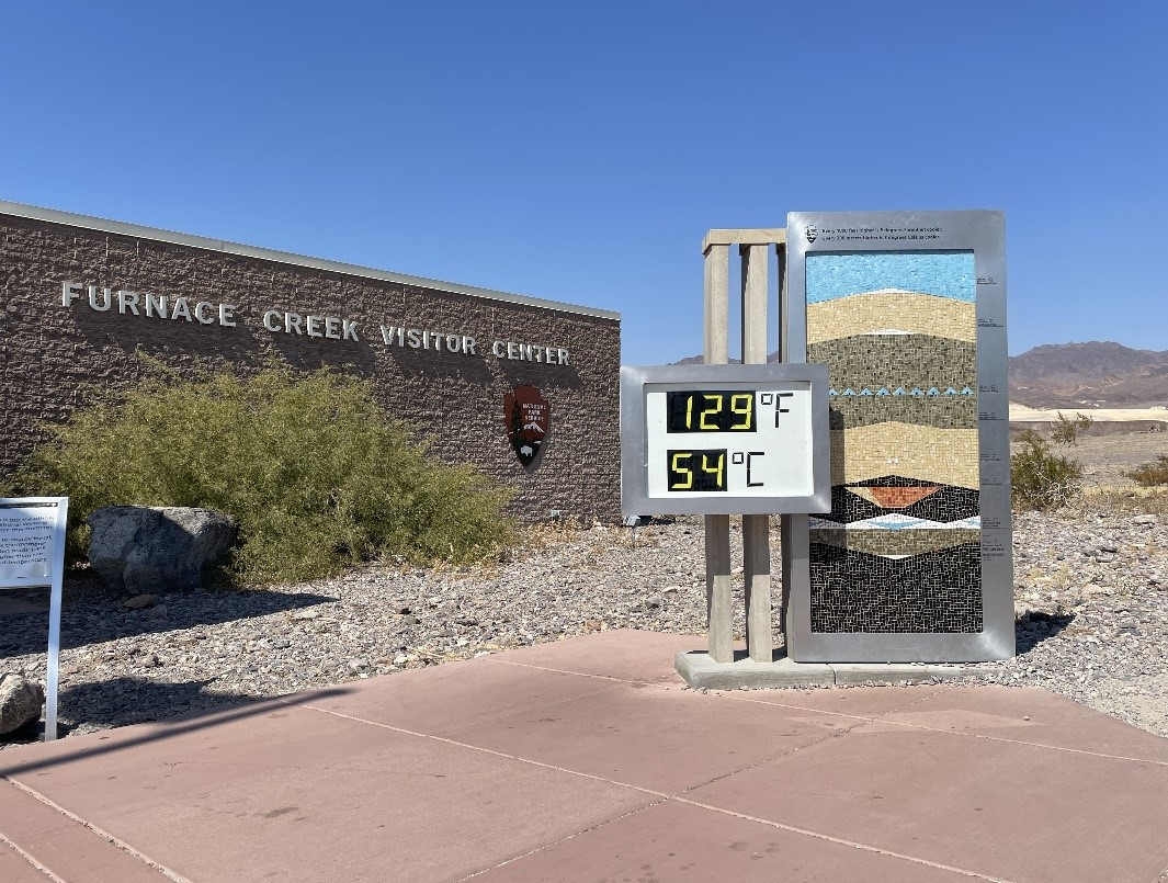 Large digital thermometer reading 129 F, 54 C in front of a brick building with the words "Furnace Creek Visitor Center" and a National Park Service arrowhead logo on the wall in a rocky desert landscape.