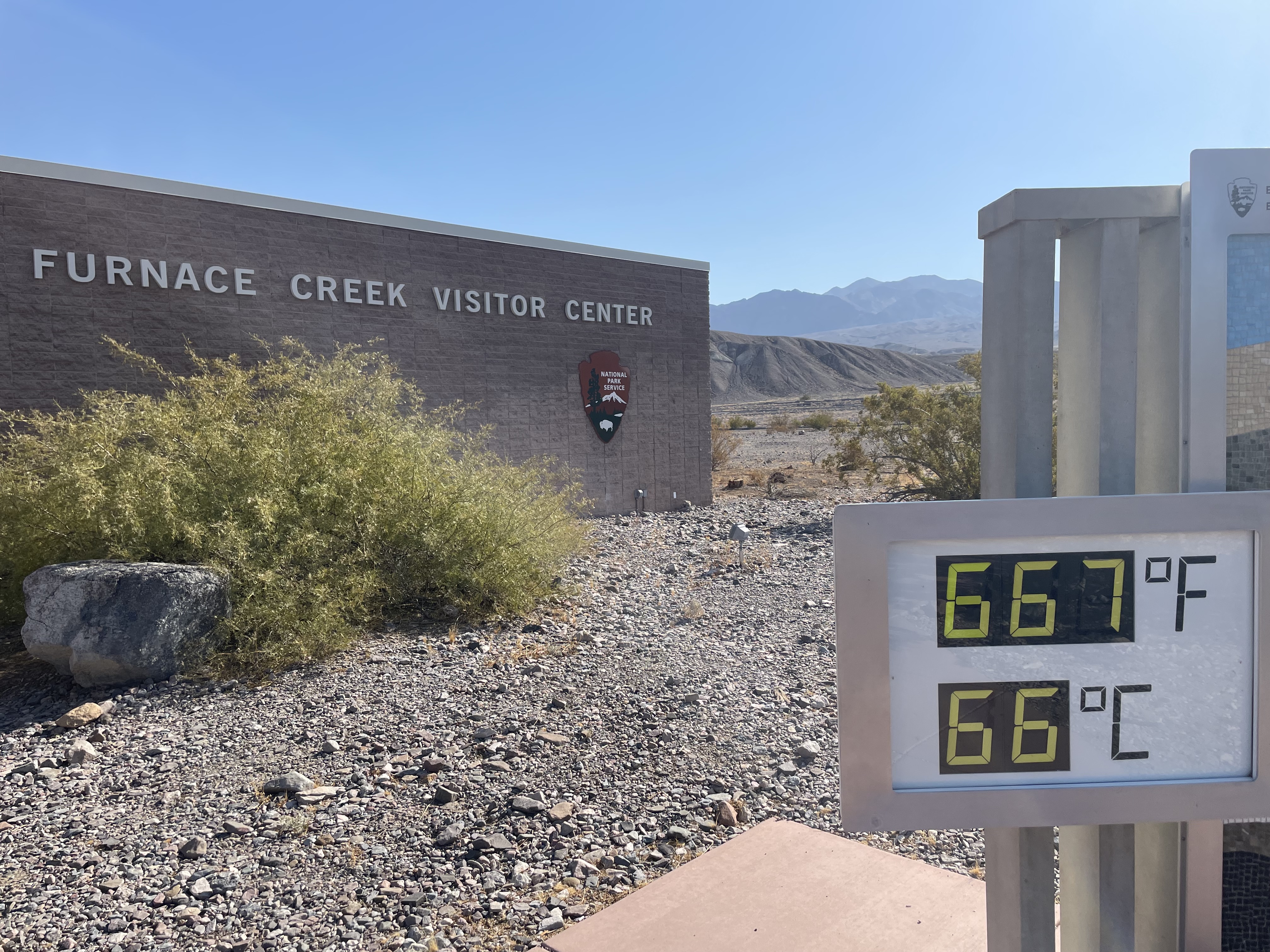 A large digital thermometer displays 667 in front of a gray building with name Furnace Creek Visitor Center.