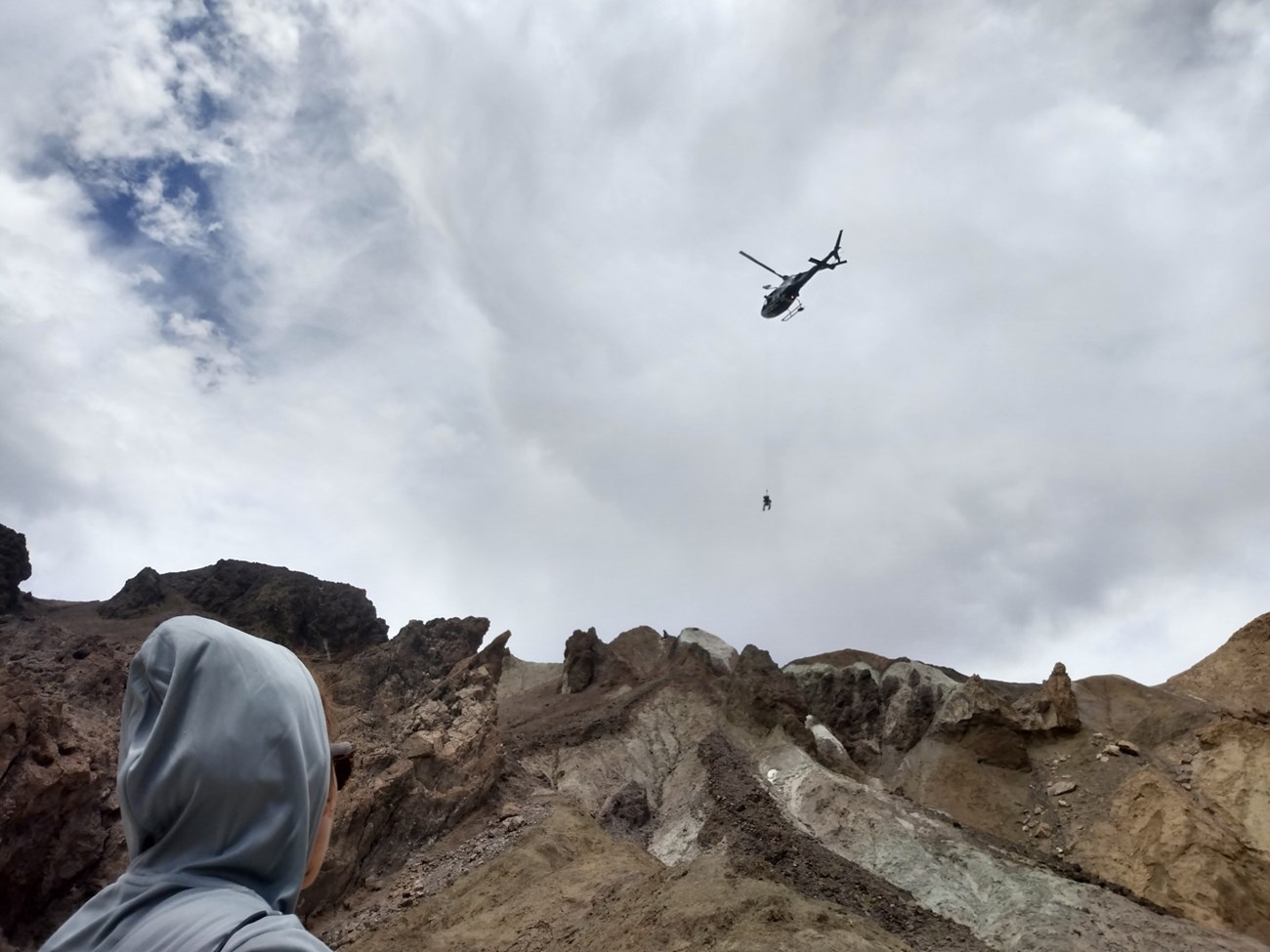 A person wearing a gray hood has their back to the camera looking up at a steep dirt and rock ridge. A person dangles from a helicopter above the ridge.