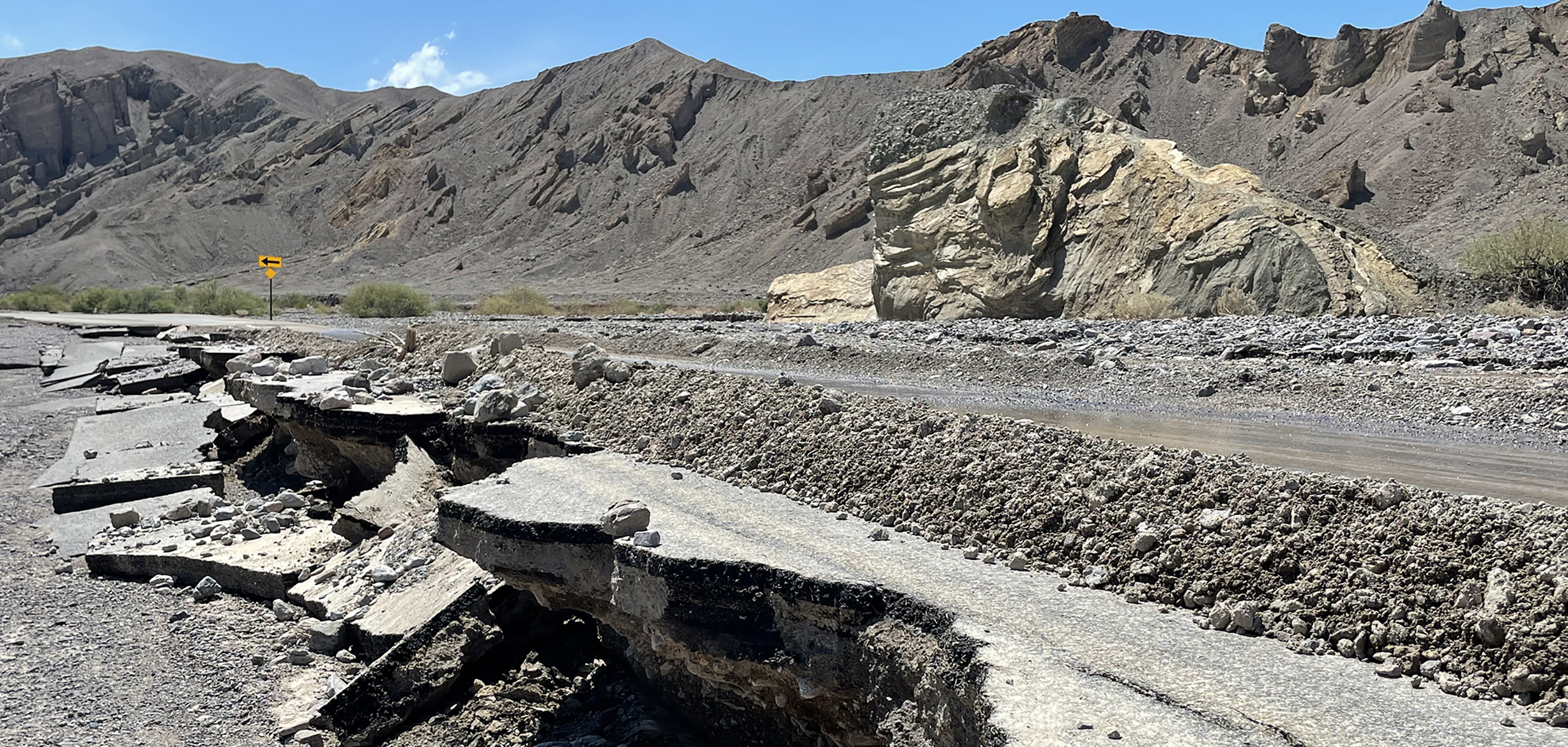 Mountain landscape frames broken pavement in the foreground of the image. The broken paved road is also covered in rocks from floods.