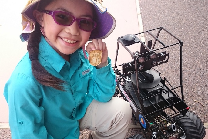 A child next to a mini Mars rover proudly shows her Junior Ranger badge
