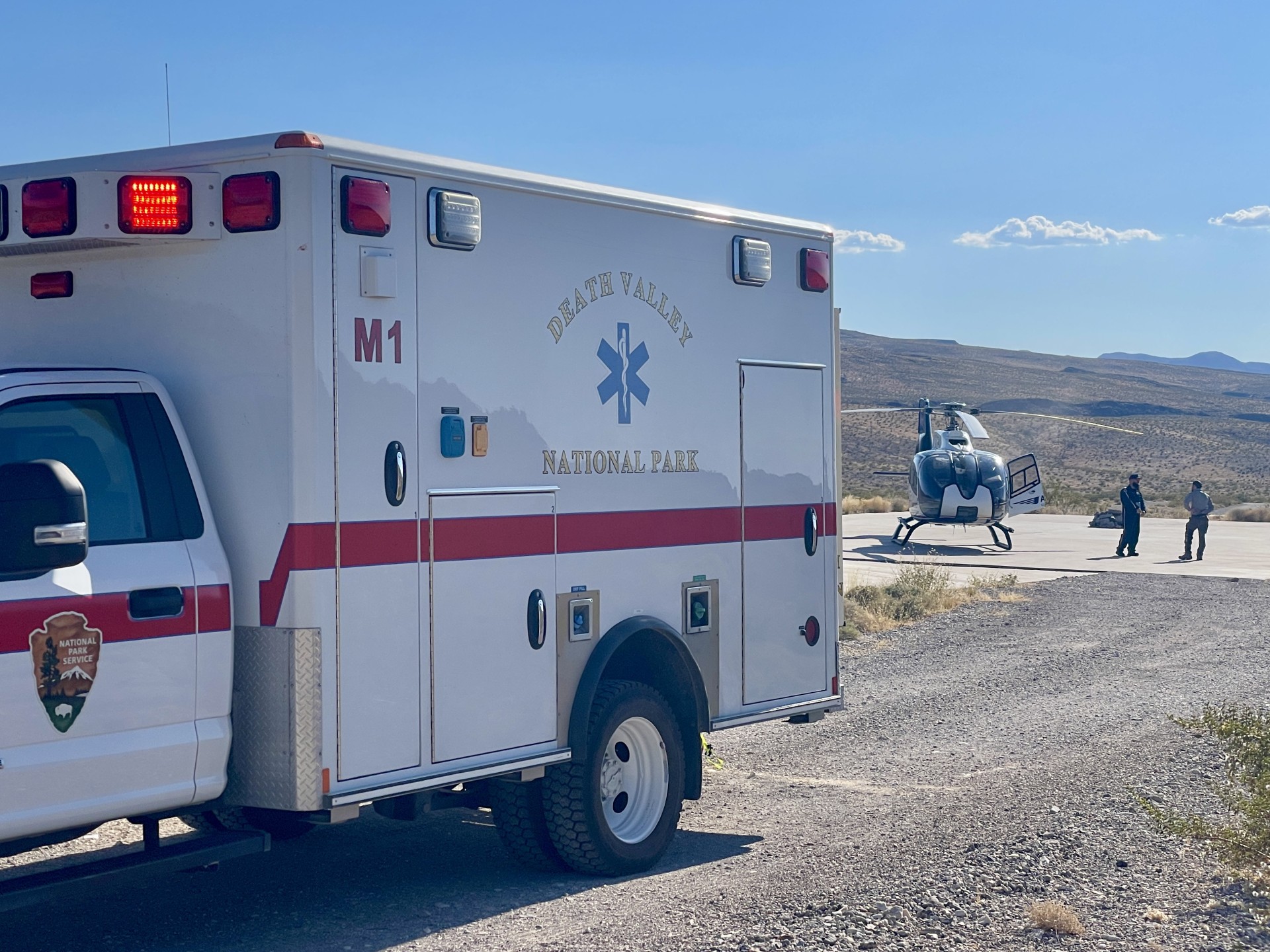 A white ambulance with a red stripe and a helicopter are in open desert landscape.
