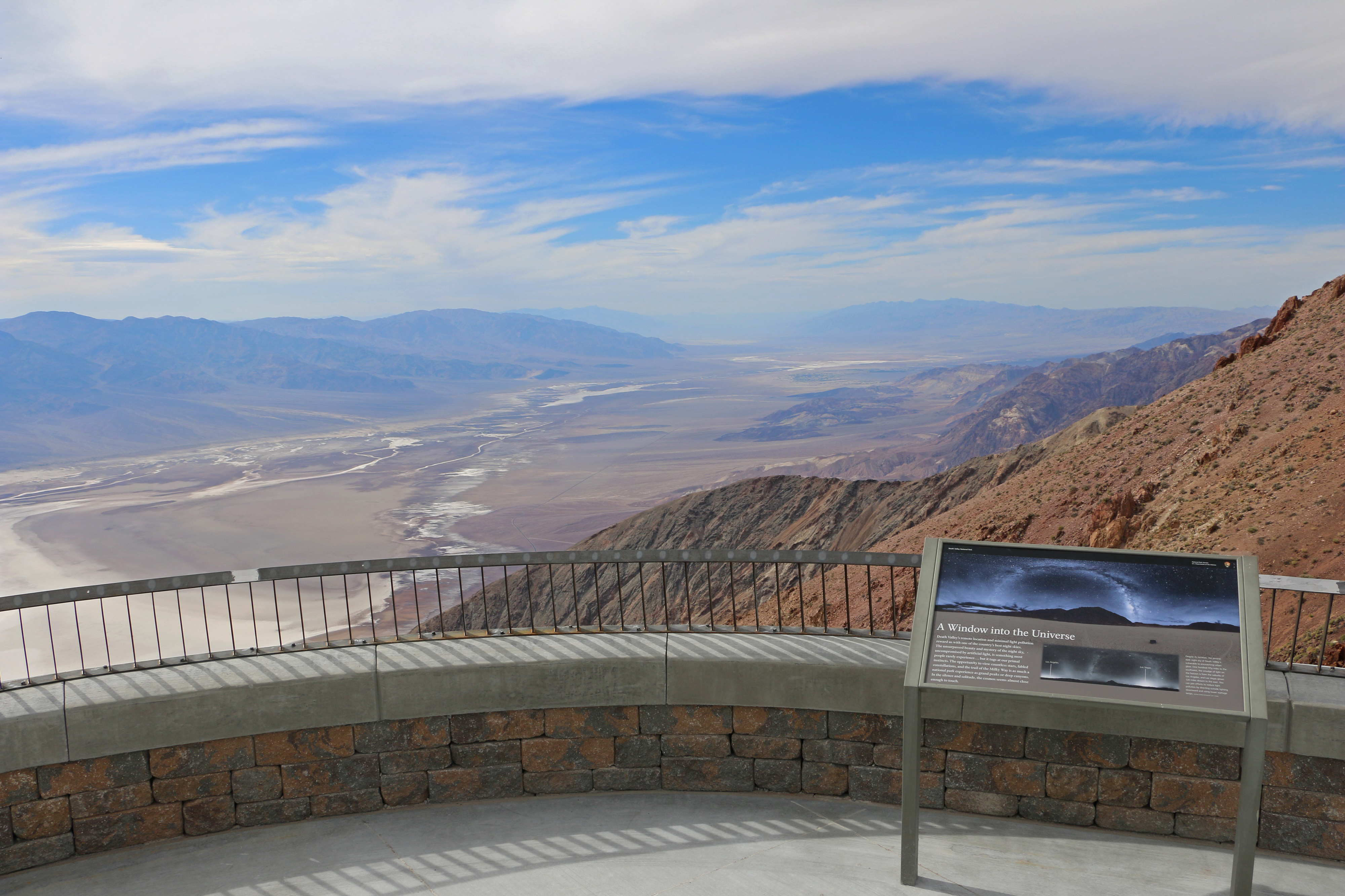 A railing and interpretive sign are in the foreground. A broad desert valley and mountains are in the distance.