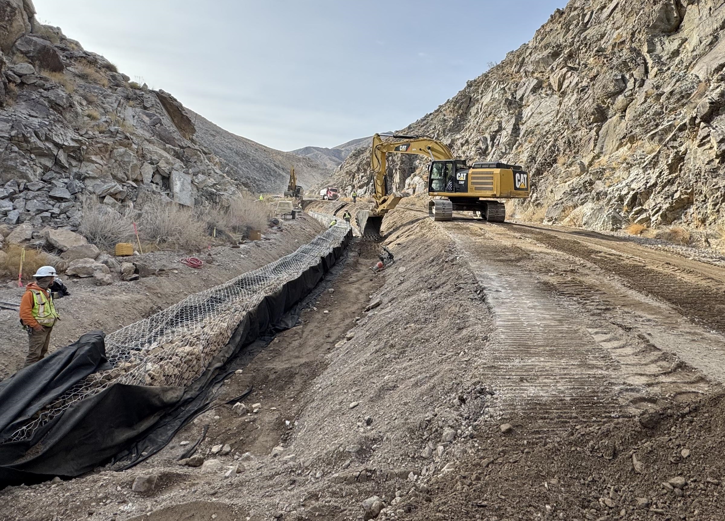 A person in a yellow vest stands next to a linear wire structure filled wth rocks. A yellow excavator and other equipment are in the background in a narrow rocky canyon.
