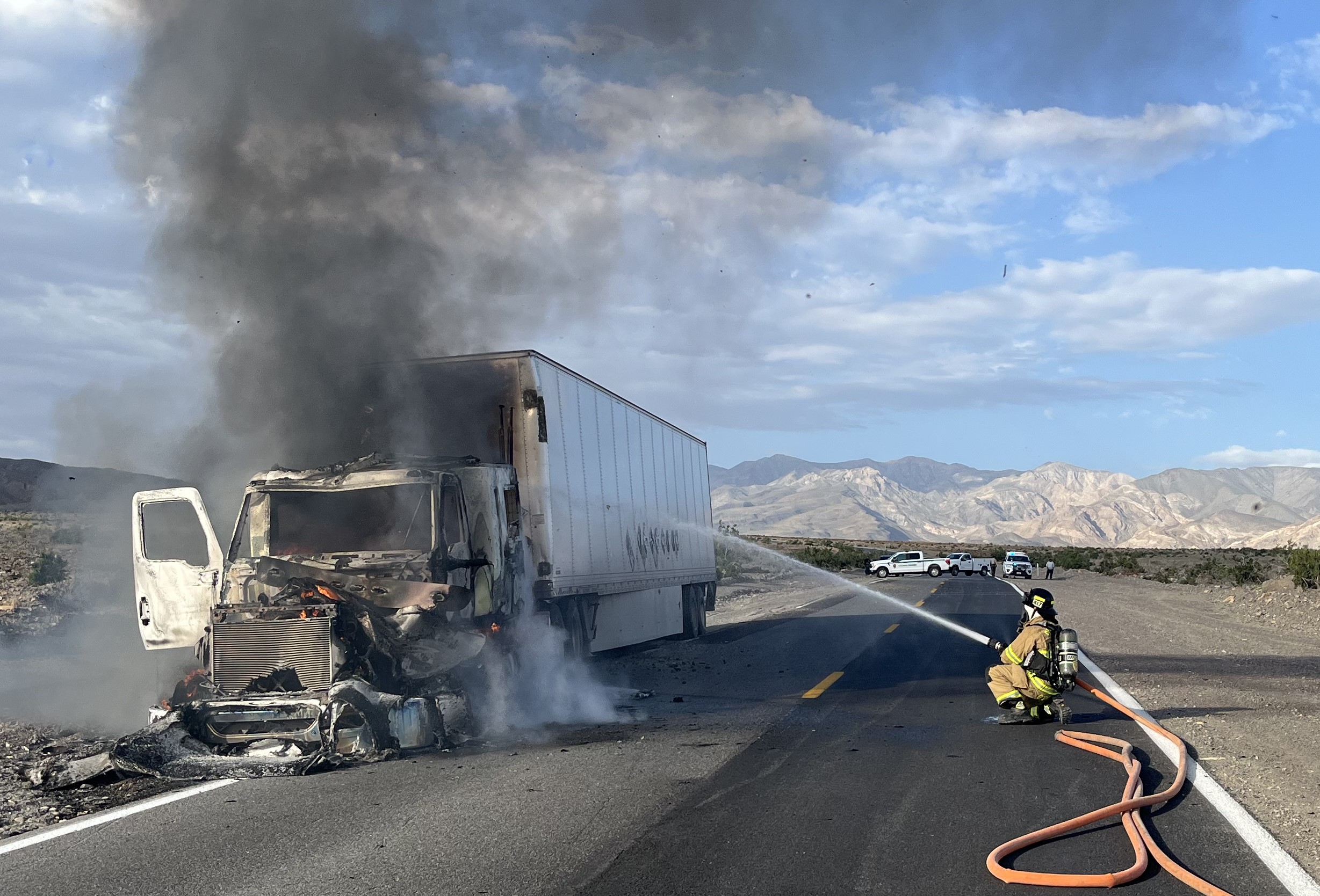 A firefighter wearing tan clothing with yellow stripes squats on the right side of the road, holding an orange firehose and spraying water at a semi truck on left side of the road. The truck's cabin is heavily damaged and smoking. The trailer looks okay.