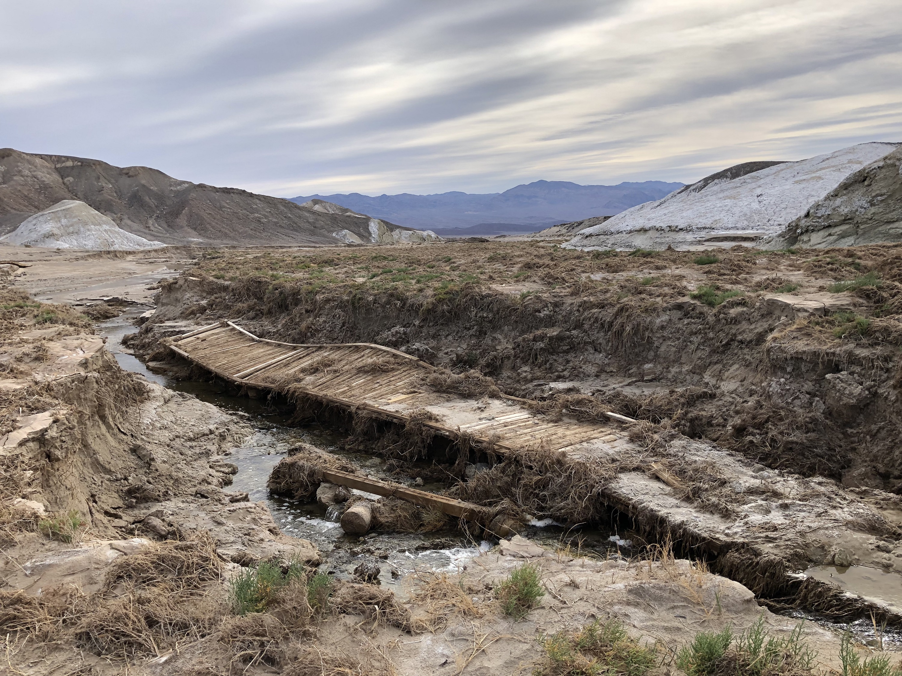 A broken and mangled segment of a wooden boardwalk is in an entrenched area between flat land with low green plants. Bare hills are in the background.