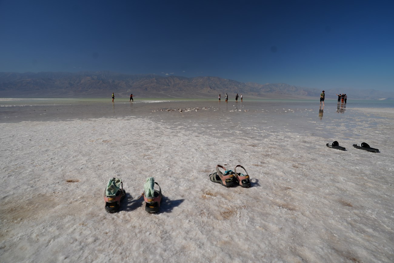 Shoes are left on a white salt flat in the foreground. People wade in shallow water in the middle distance, and stark mountains are in the background.