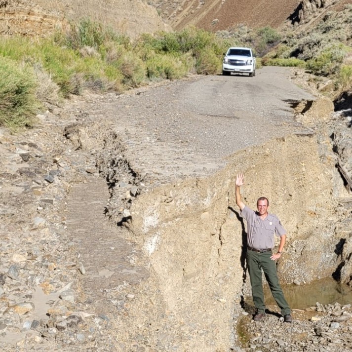 A man in green pants and gray shirt stands in front of a vertical dirt wall. Above and behind it a paved road leads between green bushes.