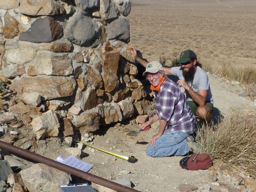 A NPS employee and a volunteer kneel by a historic stone wall with tools.