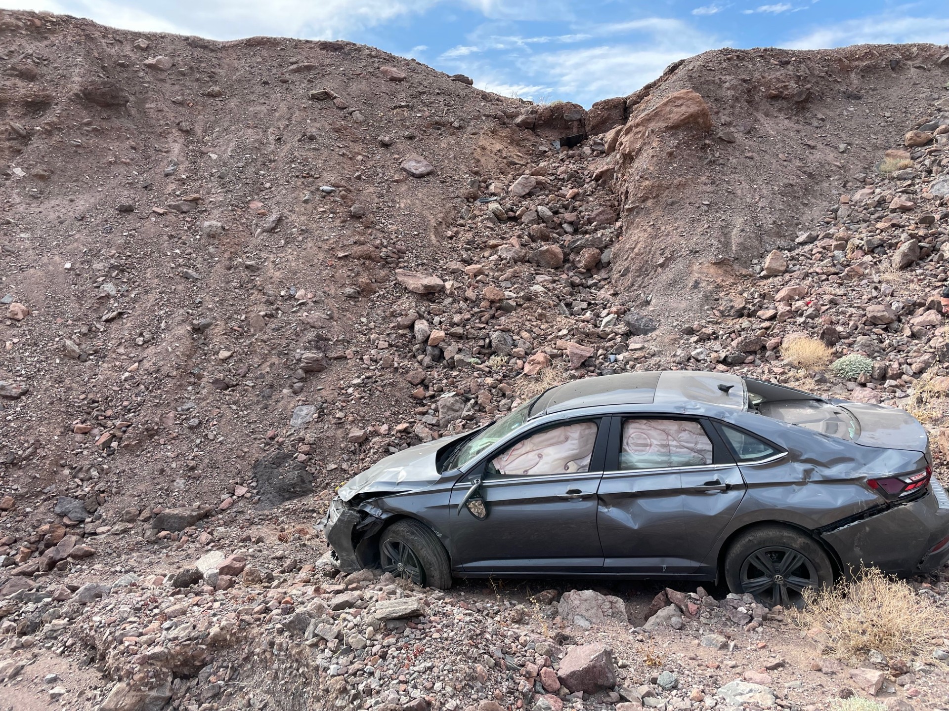 A gray car with numerous dents, a broken back window, and airbags deployed, is right-side-up at the bottom of a 20-foot-tall steep dirt and rock slope.