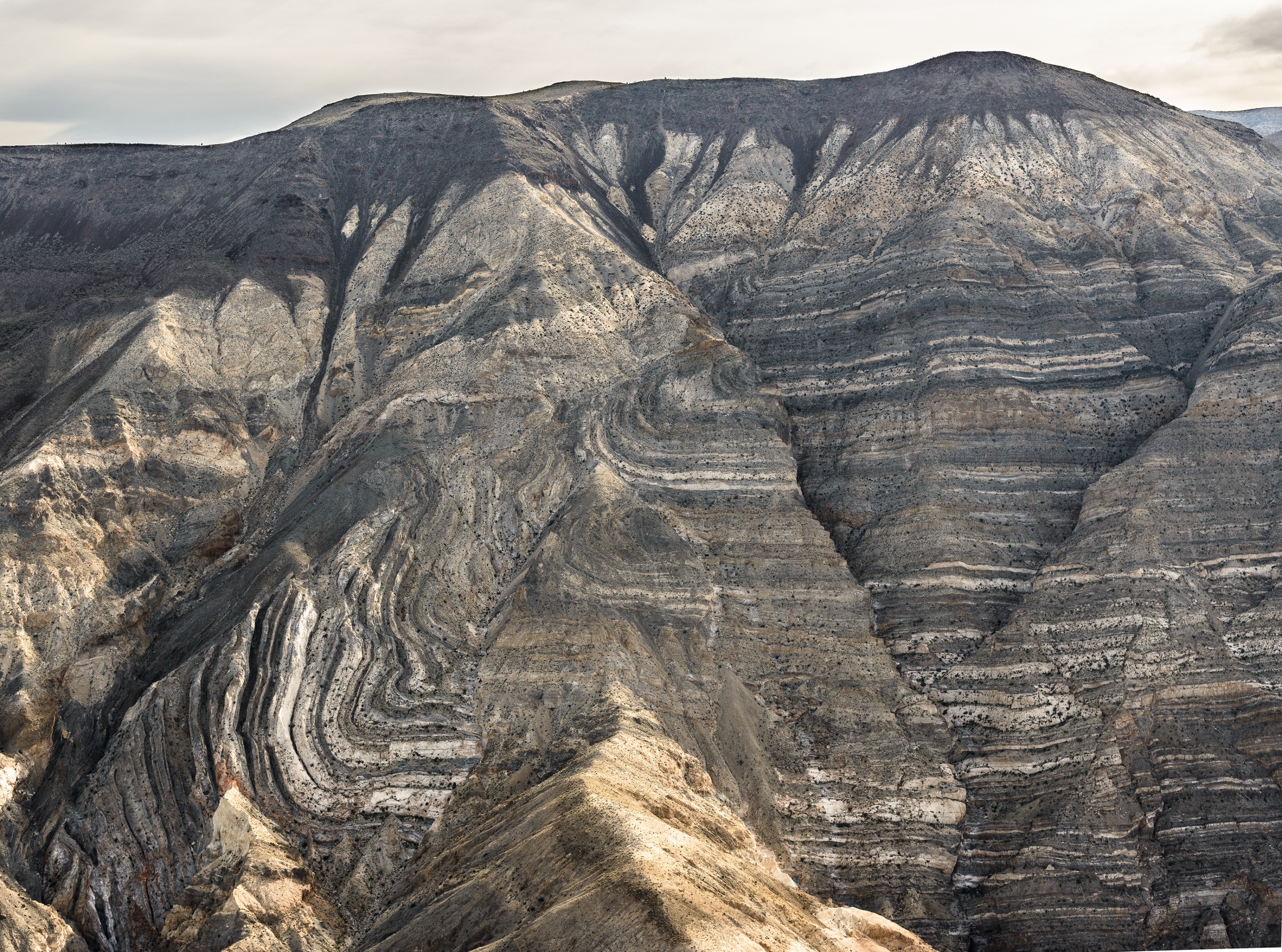 A steep mountain with no plants or trail. The mountain face is composed of twisted stripes of dark grey and almost white rock.