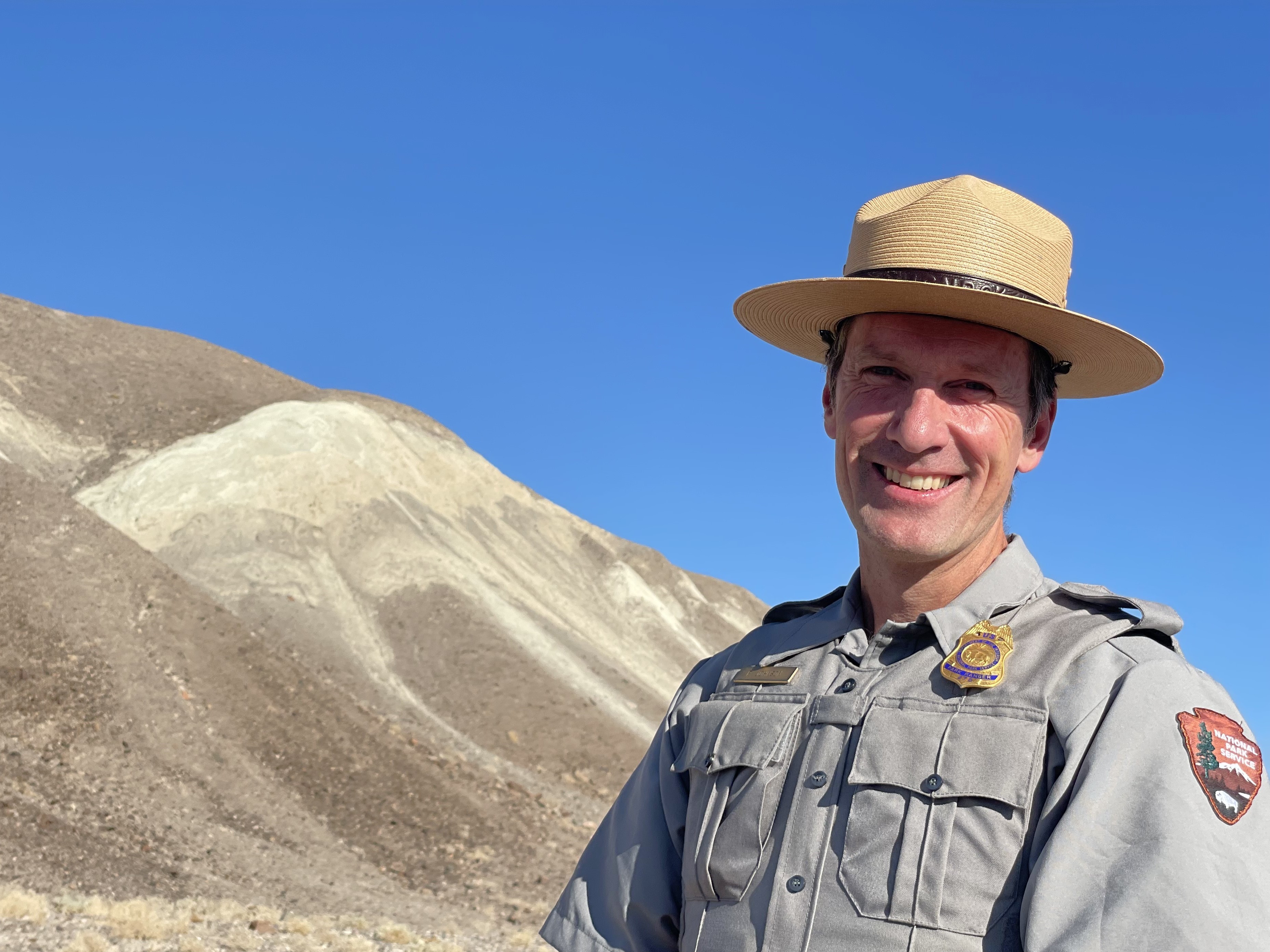 A man smiles while wearing National Park Service uniform, including a tan hat with a flat brim, gray shirt with NPS logo, and a badge. Blue sky and a brown and tan bare hill are in the background.