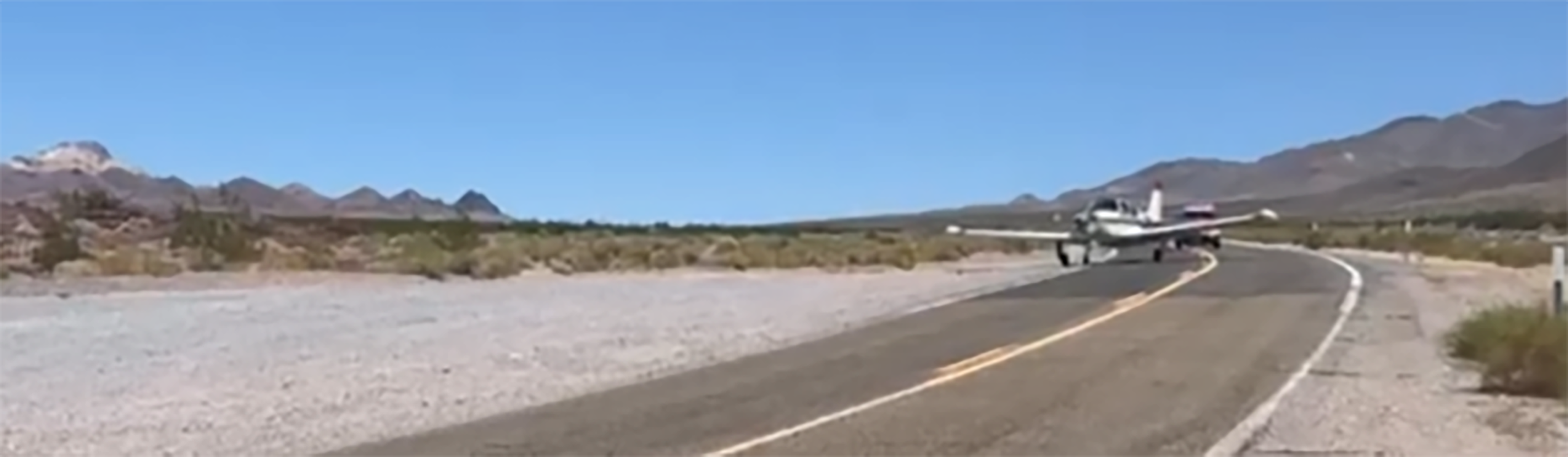 A small white airplane is on a two-lane paved road in a desert environment.