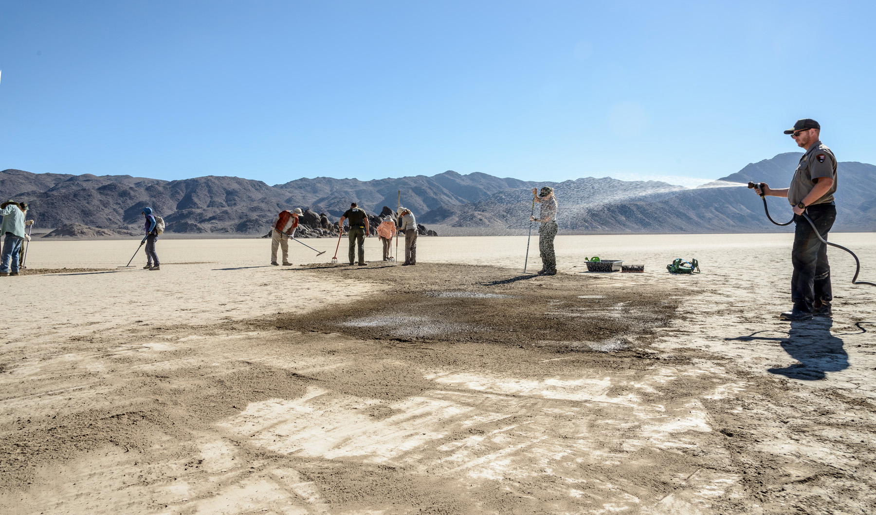 A group of people are raking and watering a large flat playa, where road tracks had been left in the dirt.