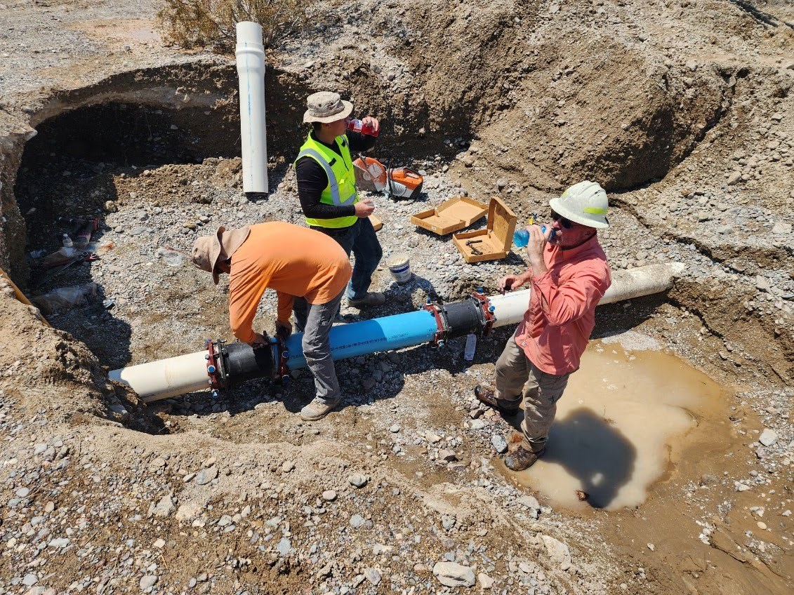 Three people stand in a puddle in a hole. Two are drinking water and one is bent over working on a pipe.