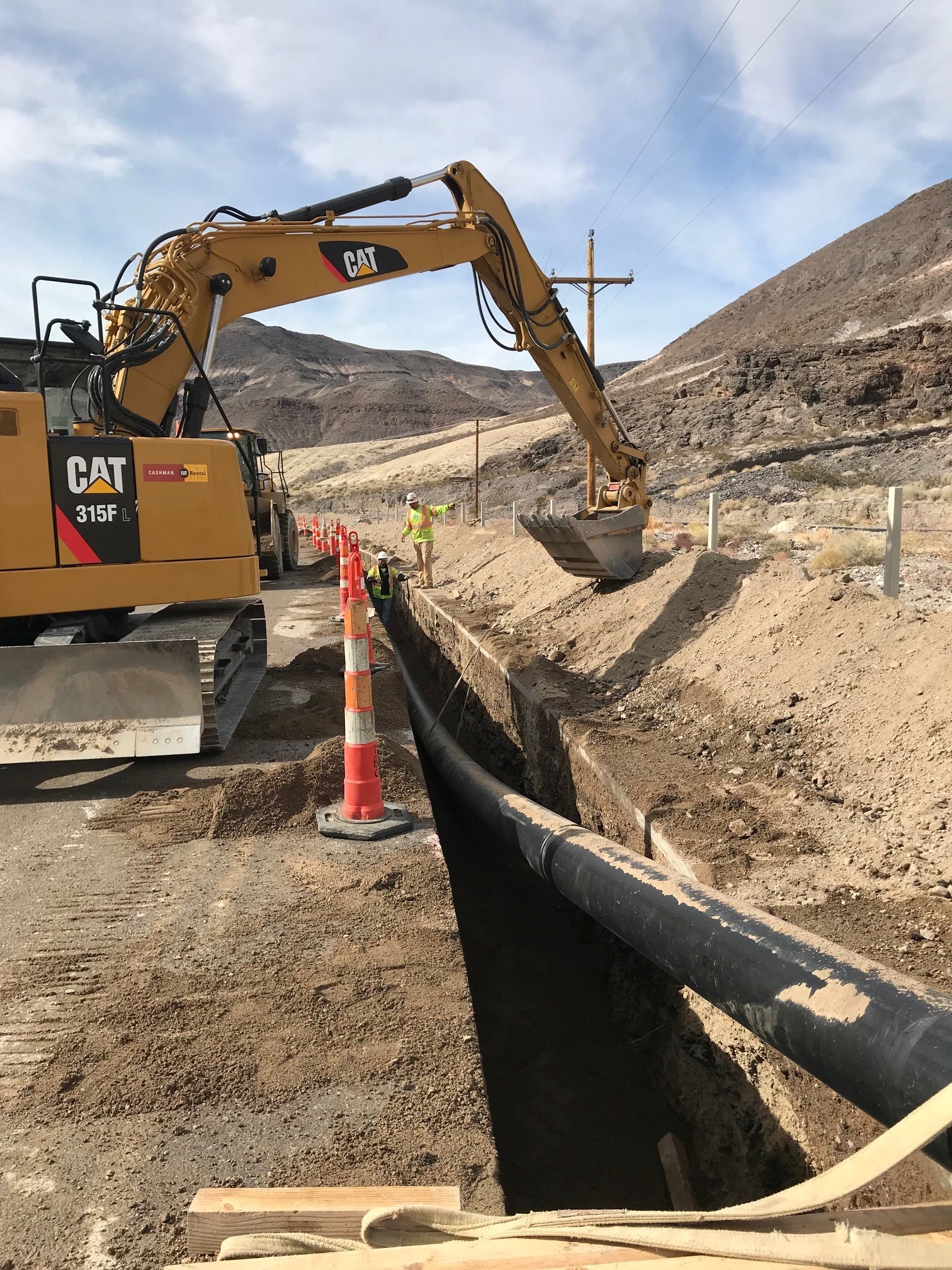 A yellow excavator is to the left of a black pipe in a trench. On the right are white fence posts.