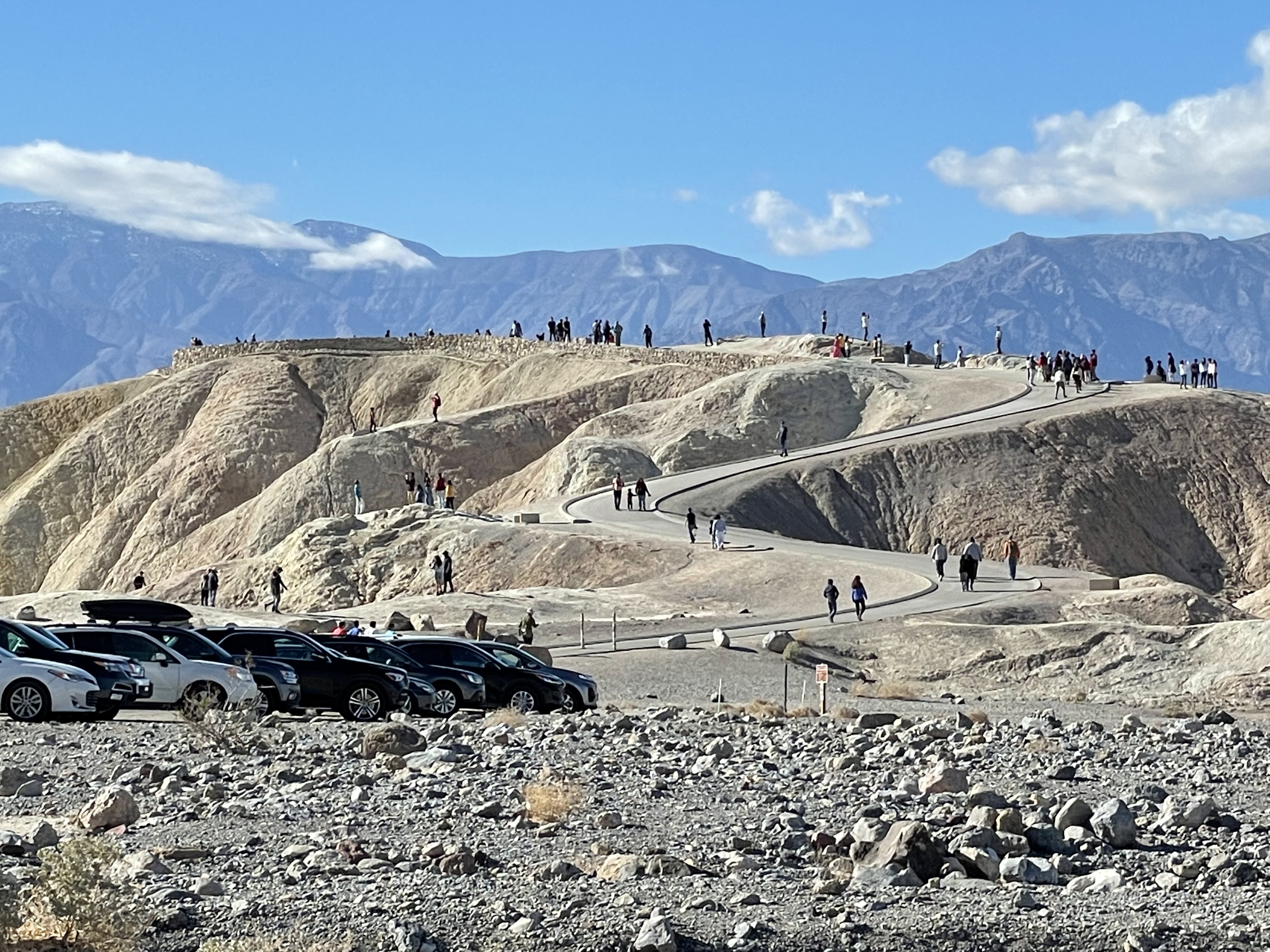 Parked cars in foreground on the left. In the middle distance people walk up a curved sidewalk on a tan-color bare hill. Dark mountains, blue skies, and a few clouds are in the distance.