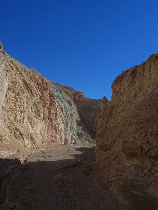 A narrow canyon with yellow, red, and green colored walls beneath a cloudy blue sky.