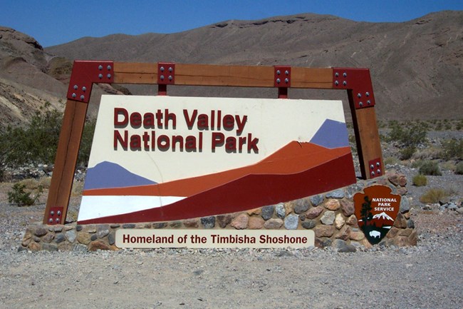 Colorful entrance sign in a desert setting with artists images of mountains and salt flats. Sign reads: Death Valley National Park Homeland of the Timbisha Shoshone. There is a wooded NPS Arrowhead logo on the stone base of the sign.
