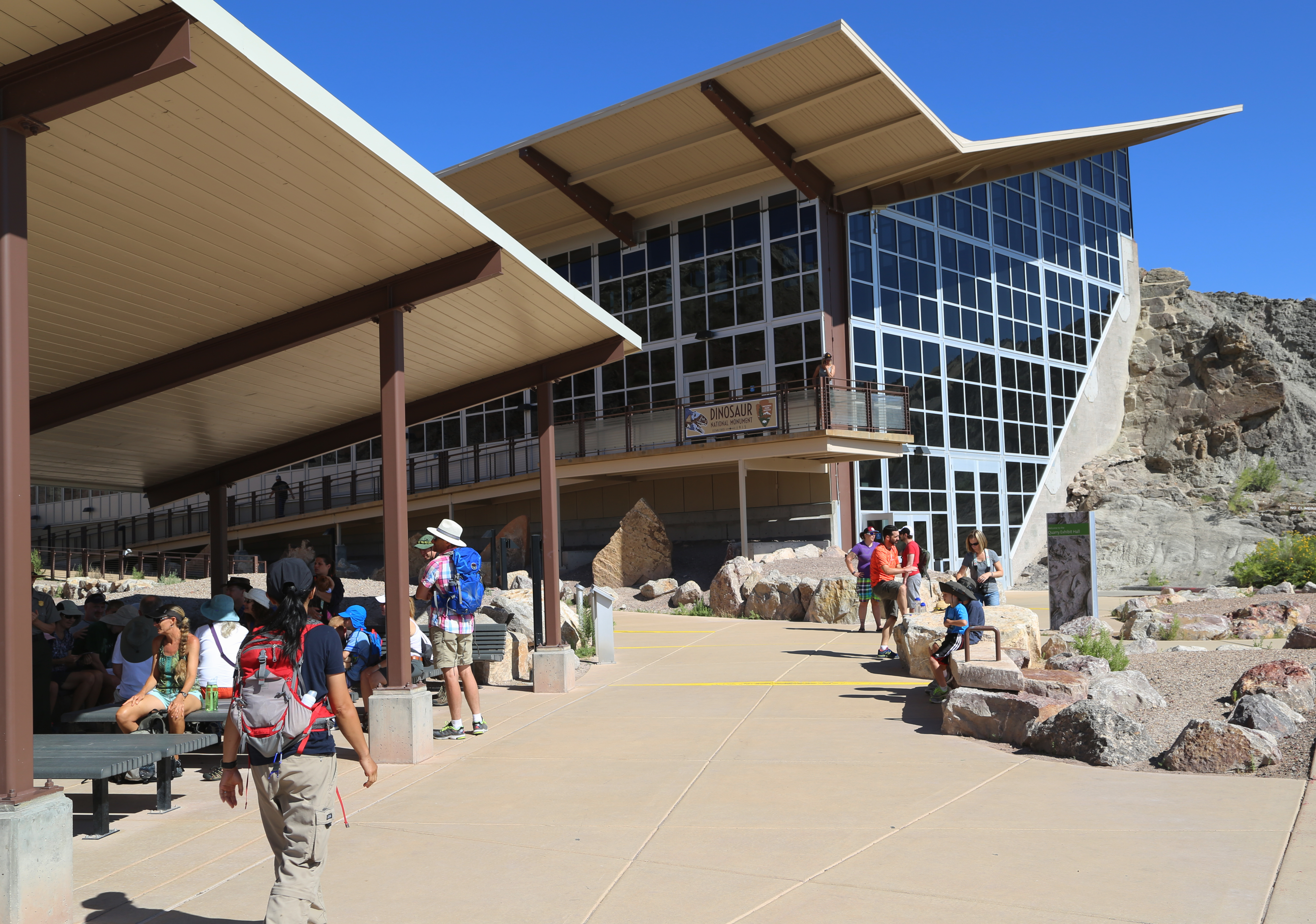 Several people walk on a sidewalk outside a building with an angled roof.