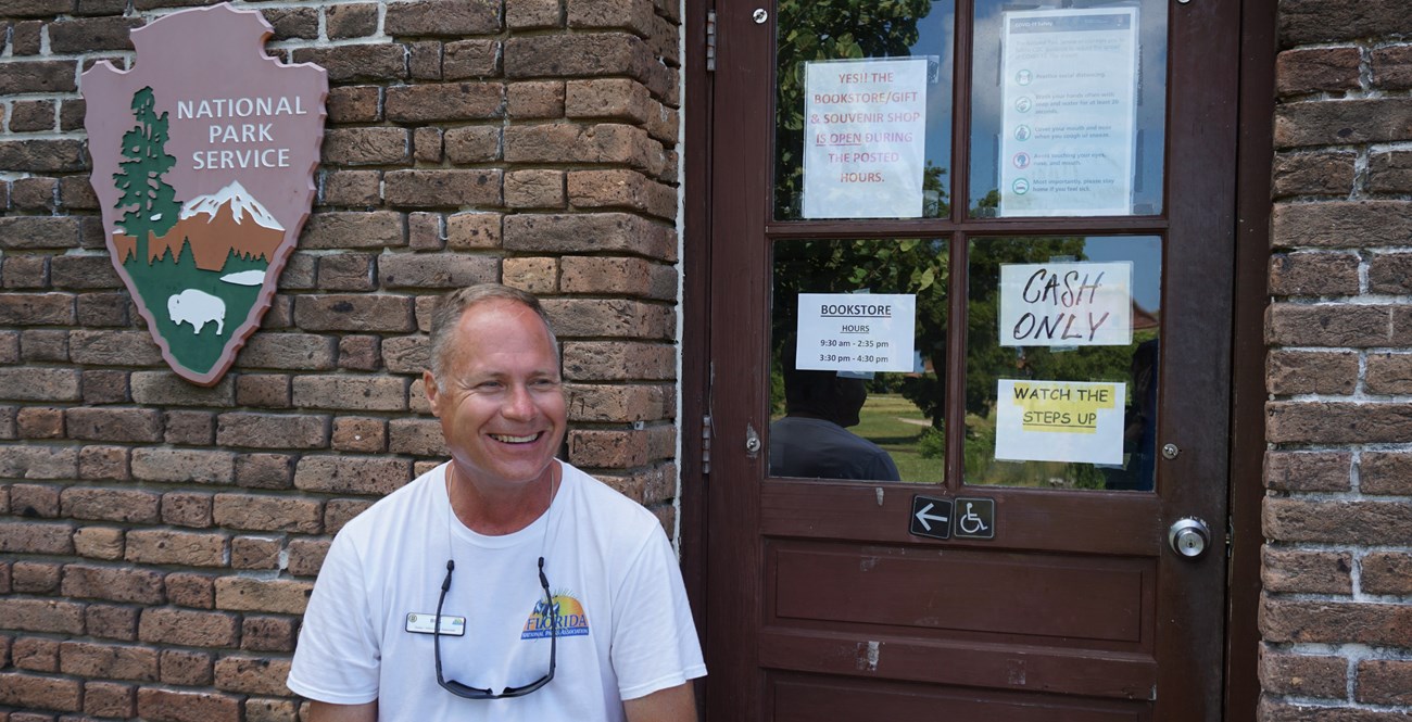 A man laughing and sitting in front of a brick building with a door and a national park service arrowhead behind him