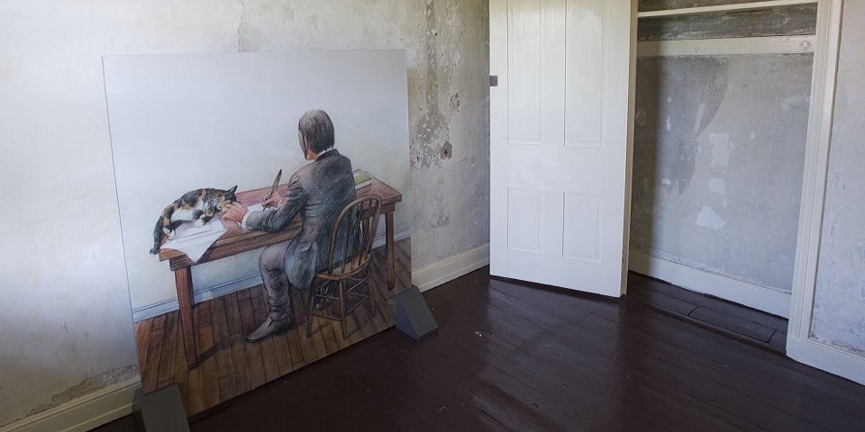 Color photo of a large illustration mounted on a wall that shows a man sitting at a desk with his back to the viewer while a calico cat reclines on the desk next to a piece of paper. " title="Color photo of an illustration showing a man sitting at a desk