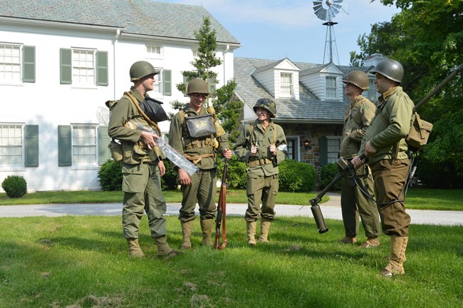 A color photograph with several men in WWII uniforms standing in front of a white brick home