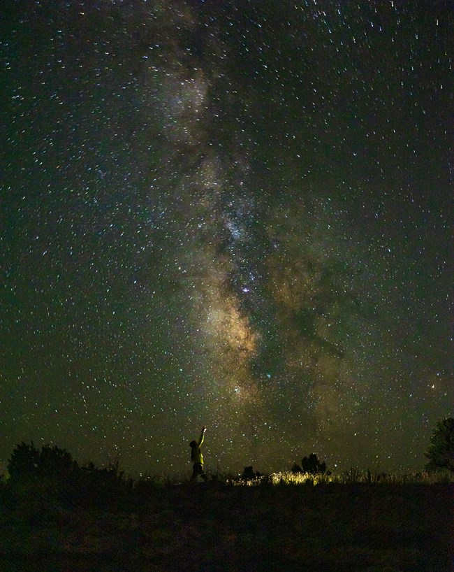 The silhouette of a person points up at the Milky Way stretching up in the night sky
